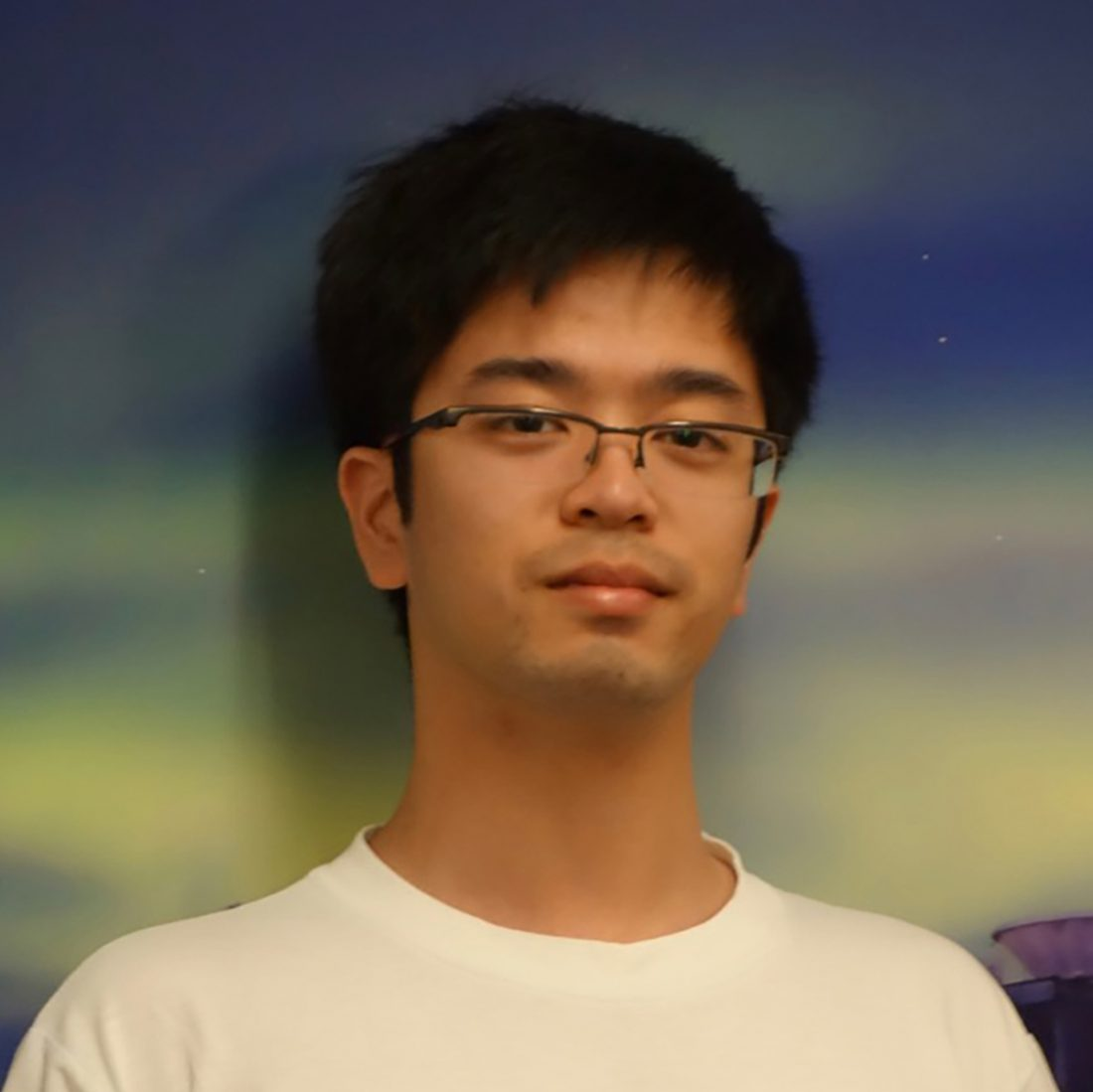 An Asian man with black hair and thin wire glasses, wearing a white tshirt and standing in front of a photo of an aurora.