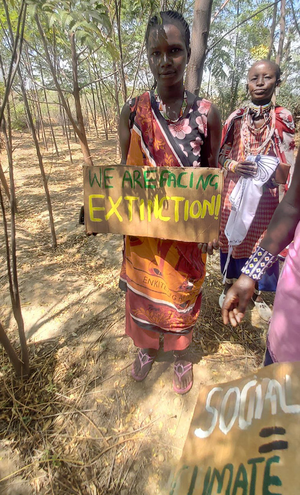 In an arid forest a Maasai woman in traditional dress holds a placard saying We Are Facing Extinction