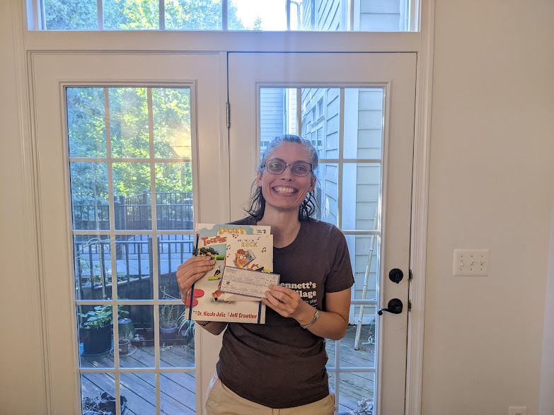 Kara, a woman with brown hair and glasses, is holding a copy of the book, Louie's Together Playground, and a check from The Able Fables for Bennett's Village.