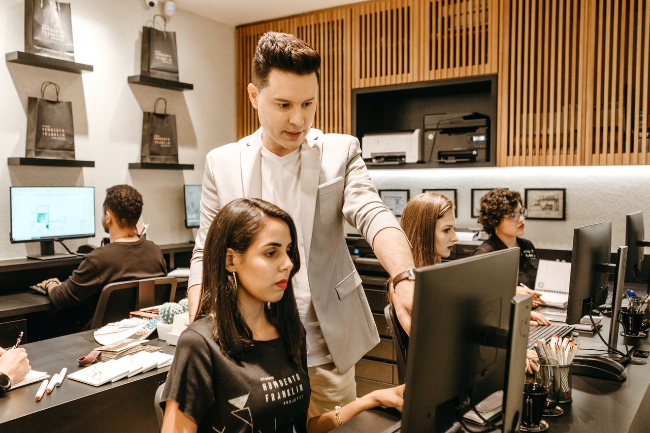 A imagem mostra um grupo de pessoas em uma sala de trabalho, com mesas e computadores. Em destaque, é possível ver uma mulher branca, de cabelo castanho liso na altura dos ombros, sentada em frente a um monitor. Ao lado esquerdo dela, há um homem branco em pé. Ele aponta para a tela do computador e parece auxiliar a mulher em alguma tarefa do trabalho. 