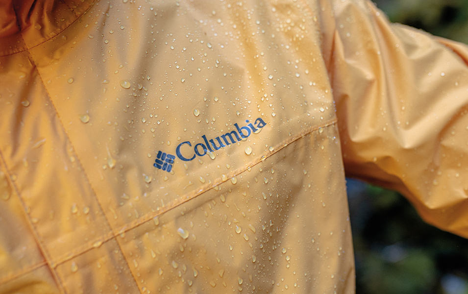 A close-up photo of the Columbia Sportswear logo on a yellow rain jacket with droplets of water all over it.