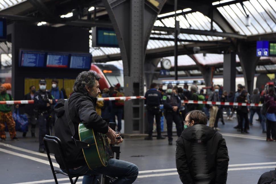 Rebels kneel and look at a rebel with a guitar in a cleared space of the station with tape and security all around it.