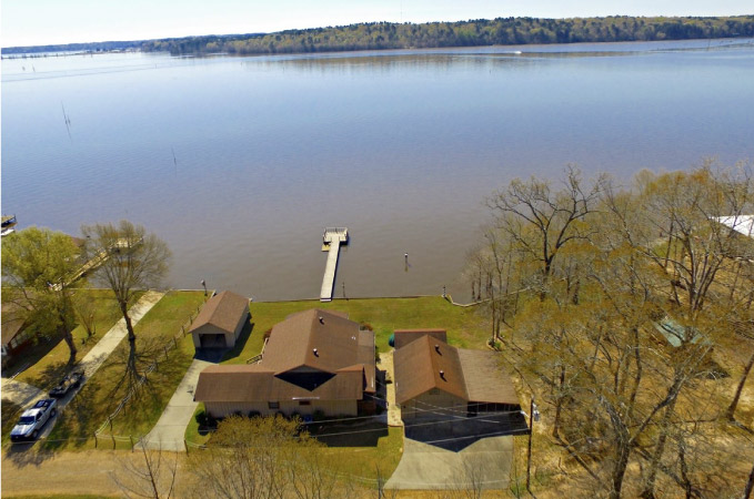 A large, lakefront home in Farmerville, LA, on Lake D'Arbonne. There’s a private dock running out into the lake and the land across the water is lush with trees.