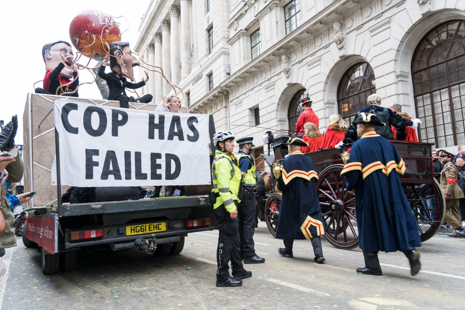 A float with puppets of political figures and a red hot world and a banner that says COP has failed. Police surround it while the Lord Mayor's carriage passes with men in pompous traditional uniforms inside