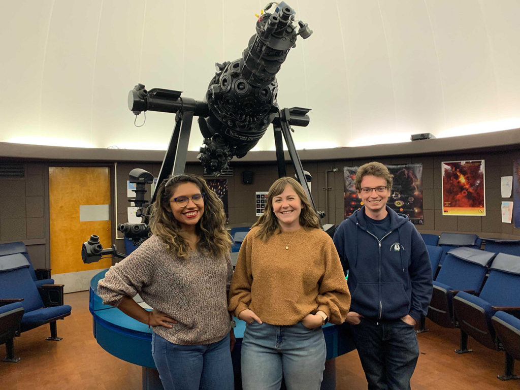 Three people (a Hispanic woman with long brown hair, a white woman with long light brown hair, and a white man with curly brown hair) standing in front of a planetarium projector.