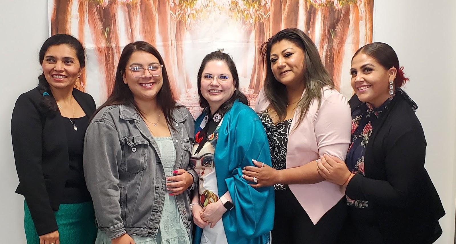 A group of five women in professional business attire stand close together at ninety degrees to the camera. They are smiling and holding each other's elbows in a friendly way.