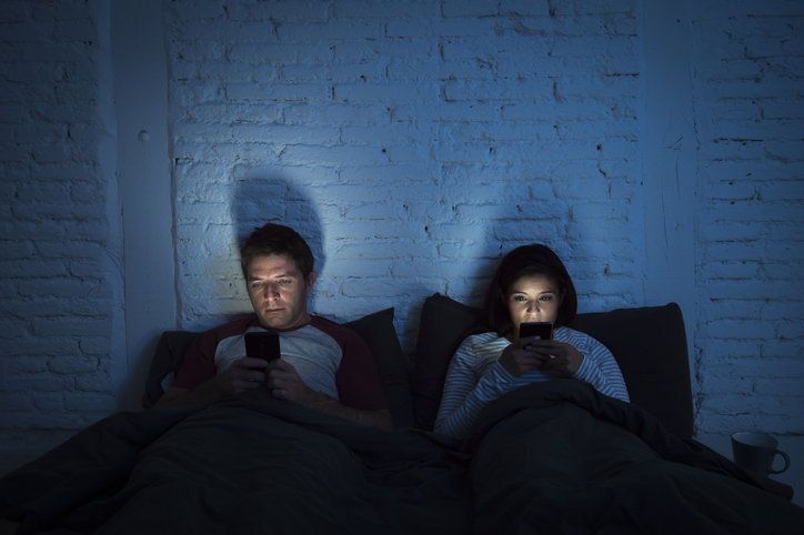 A couple in bed with the lights off using their phones. Turn off electric devices before bed