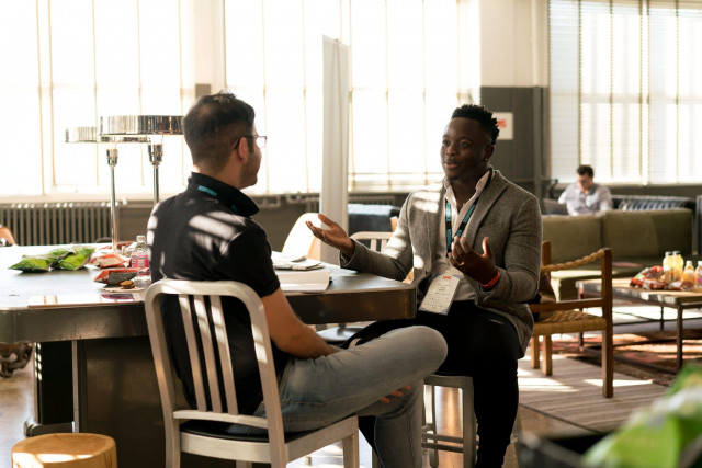 Imagem de duas pessoas conversando frente a frente, sentadas em cadeiras, em um café. Enquanto um homem de terno fala e gesticula, o outro presta atenção nas suas palavras. A feição de satisfação de ambos representa a importância da experiência do cliente