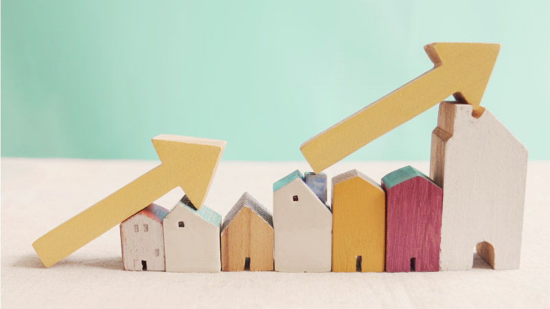 A line of small wooden blocks in the shape of houses, each one bigger than the last, with wooden arrows leaning against them pointing up and to the right — signifying elevated home prices due to inflation.