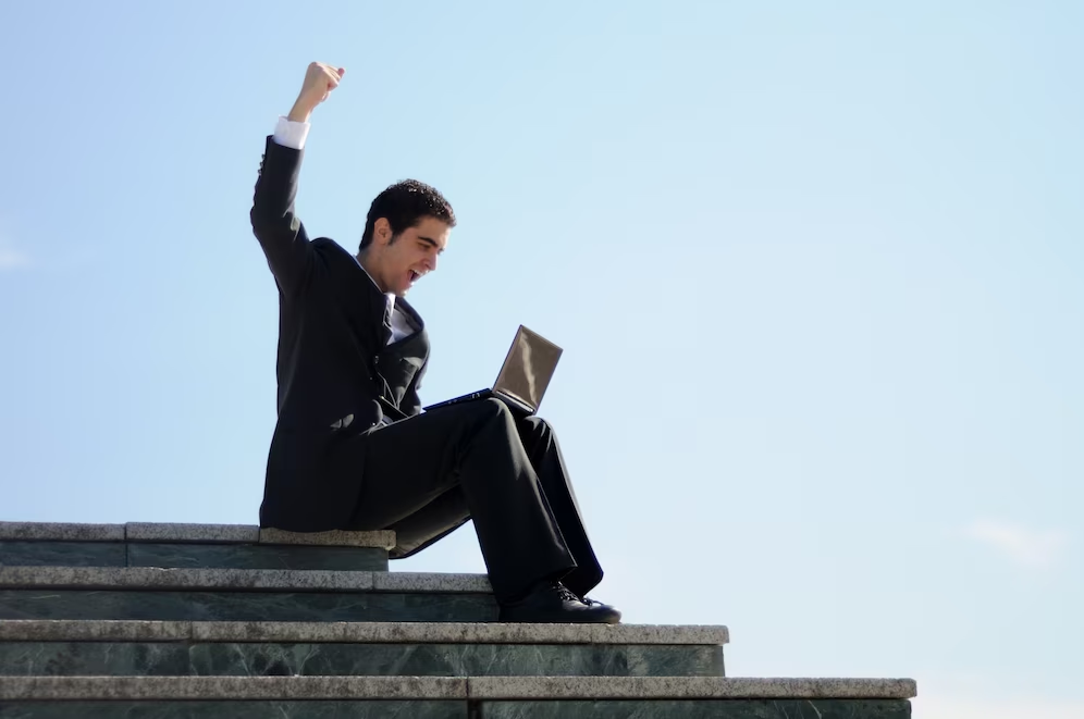 A man sitting on outdoor stairs, using a laptop, with raised hands