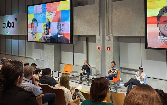 Foto da apresentação "O futuro do entretenimento", no Fast Company Innovation Festival Brasil 2022. Ao centro, está Eduardo Tracanella, um homem branco, careca e vestido com uma camisa preta e jeans; Duda Magalhães, homem branco, de cabelos curtos e encaracolados, de camisa xadres preta e branca, e calça jeans; e Marcelo S2, um homem negro, de boné preto, camiseta branca e calça preta. Eles estão de frente para a plateia, ao fundo está uma parede de concreto e acima está um telão com as fotos dos participantes com fundo colorido.