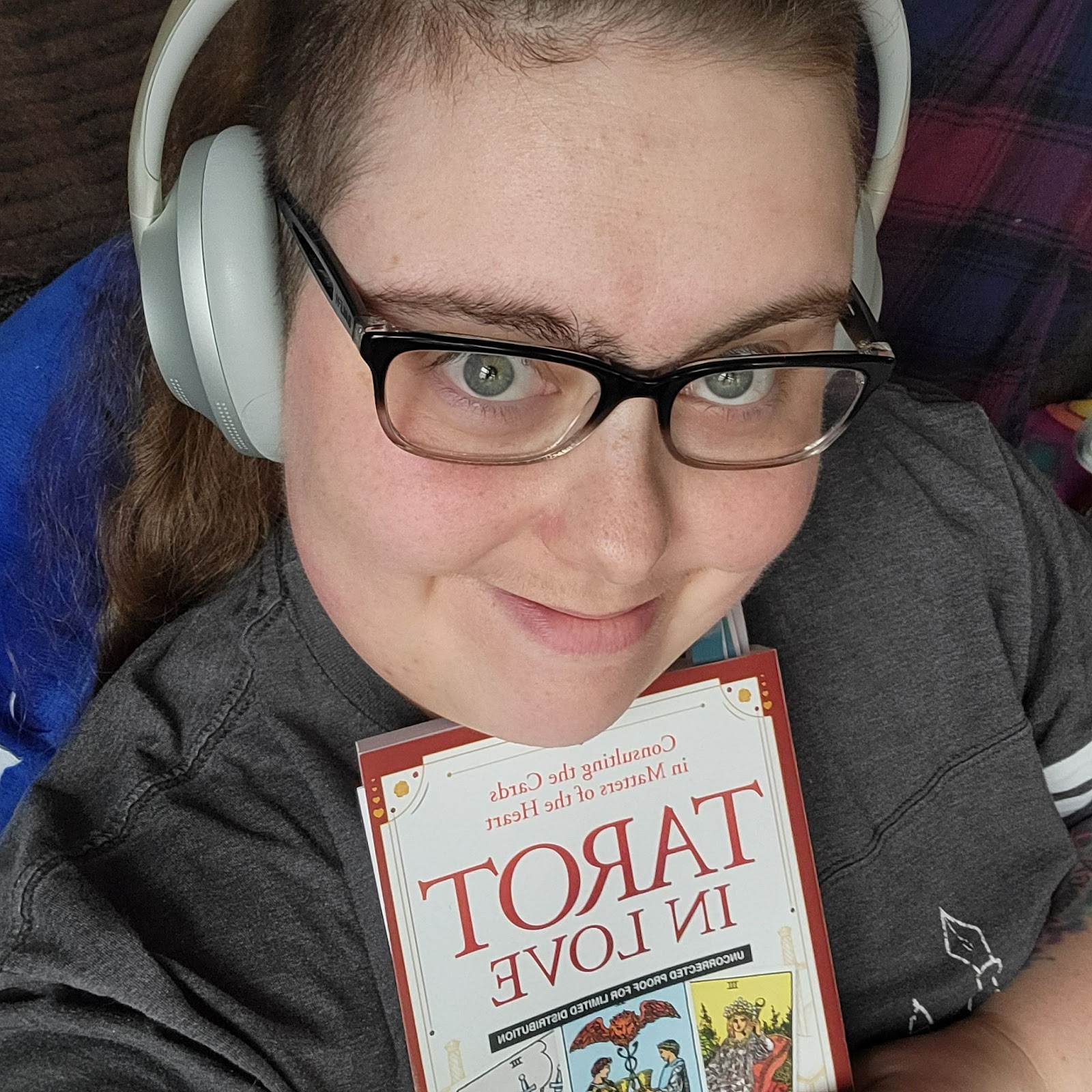 Image description: Ru-Lee Story, a nonbinary person with blue-green eyes and spectacles, smiles at the camera while holding an advanced reader copy of Tarot in Love by Elliot Adam.