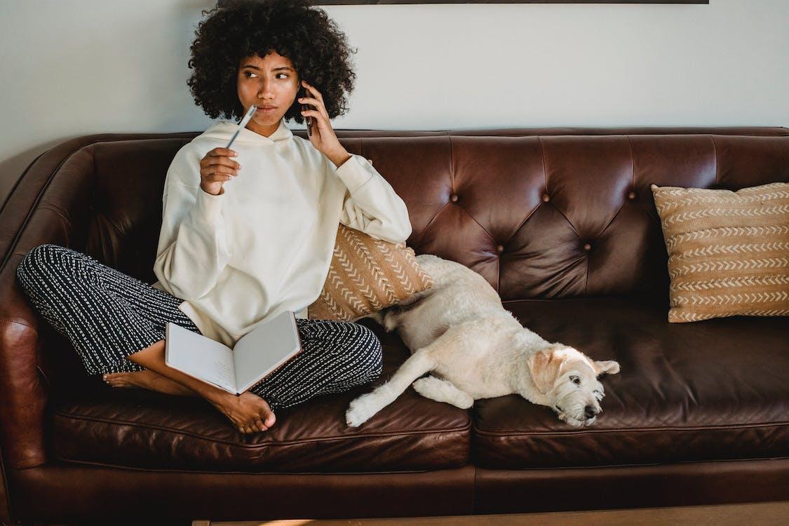 Free Thoughtful black woman with notebook and dog on leather sofa Stock Photo