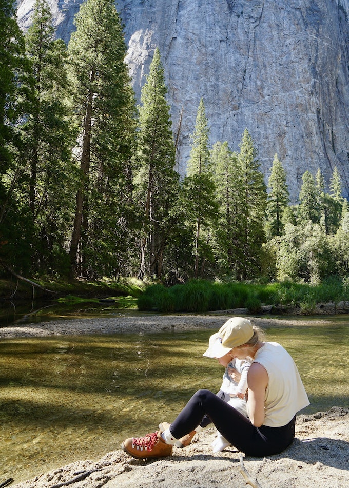 A person is sitting next to a river playing with a toddler. The river is lined with trees and there's a tall rock wall in the background.