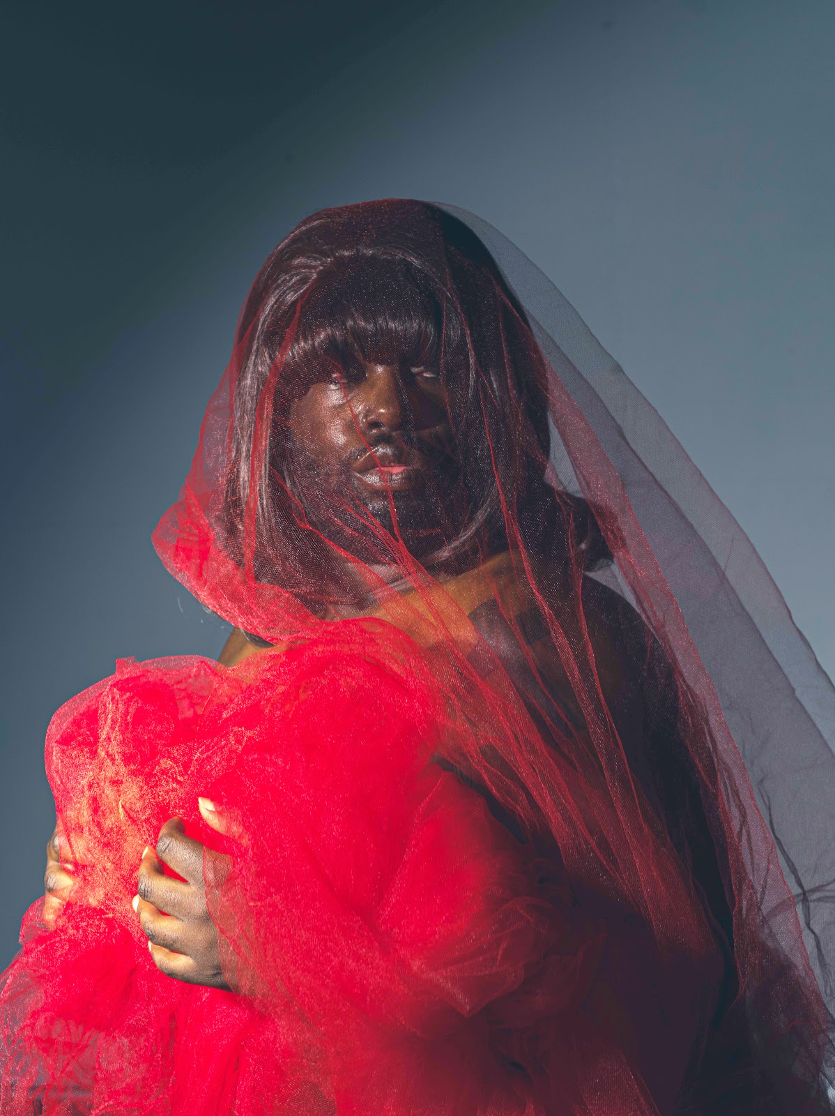 Image right: Shabez Jamal, Anita in Red, 2023. A portrait of Anita against a dark gray and gradient background, glancing to the right, beyond the image frame. They are wrapped in bright red tulle with a thin layer covering their head like a veil. Photo courtesy of Shabez Jamal.