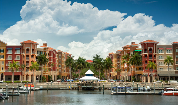 A downtown shopping area and marina in Naples, Florida. The buildings are painted peach and coral hues and there are fluffy white clouds in the sky.