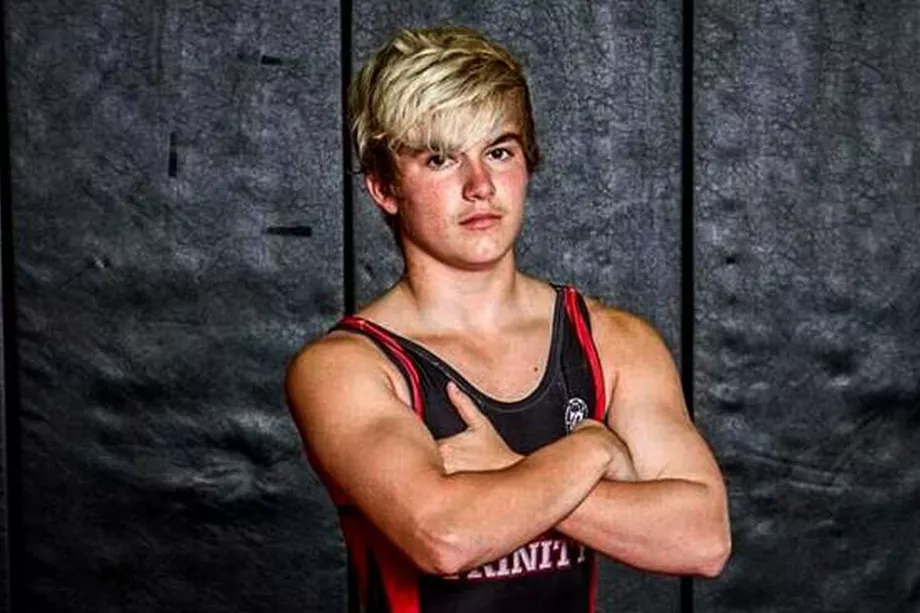 A young man with a shock of dyed blond hair and pale skin slightly flushed from exertion stands with his arms crossed, looking straight into the camera, against a black background wall. He wears a black school wrestling tank with red trim and white letters that say "TRINITY," the name of his school. 