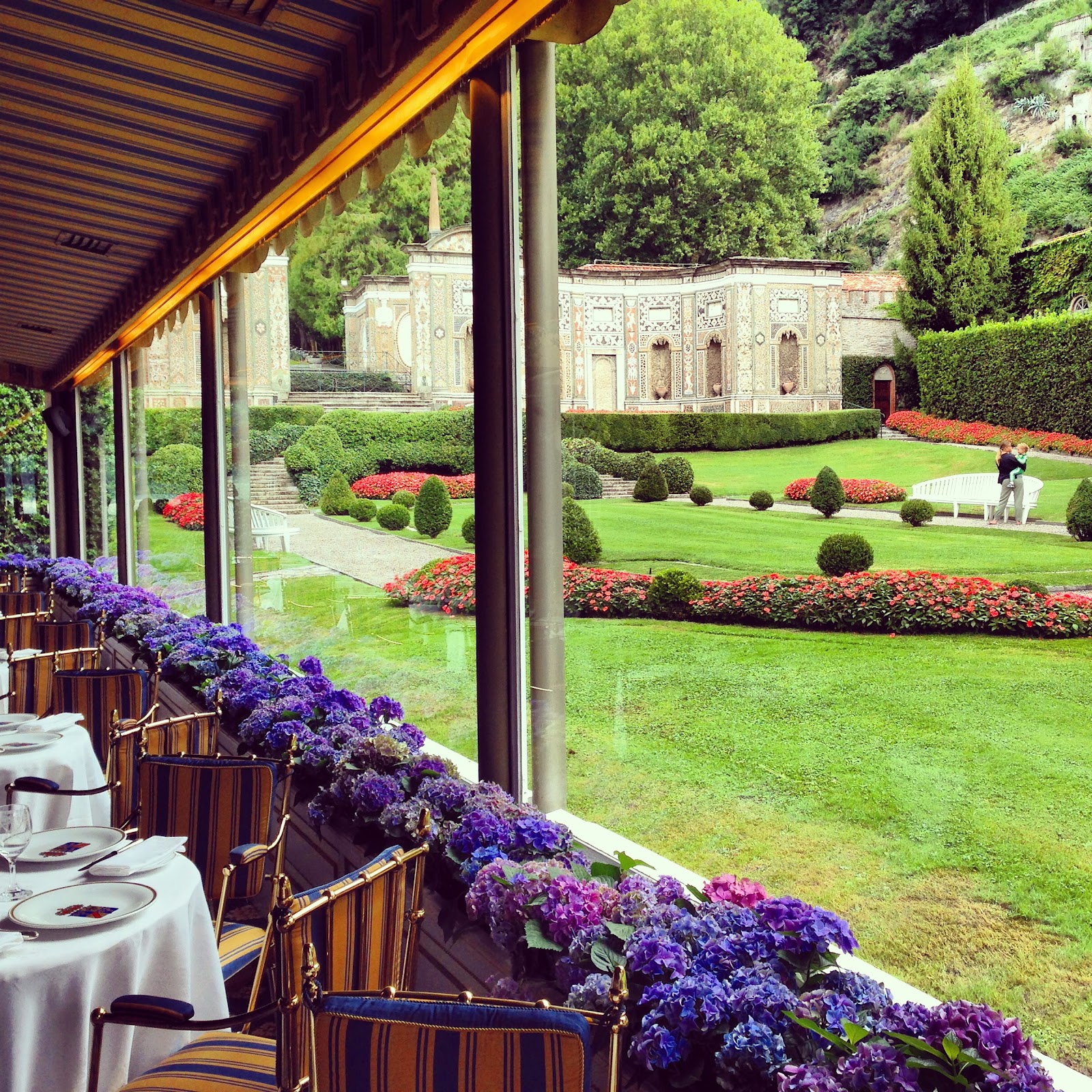 villa d'este restaurant. View the villa's beautiful garden while dining in the terrace. Empty, decorated tables, windows lined with purple flowers and park with tourist on bench.