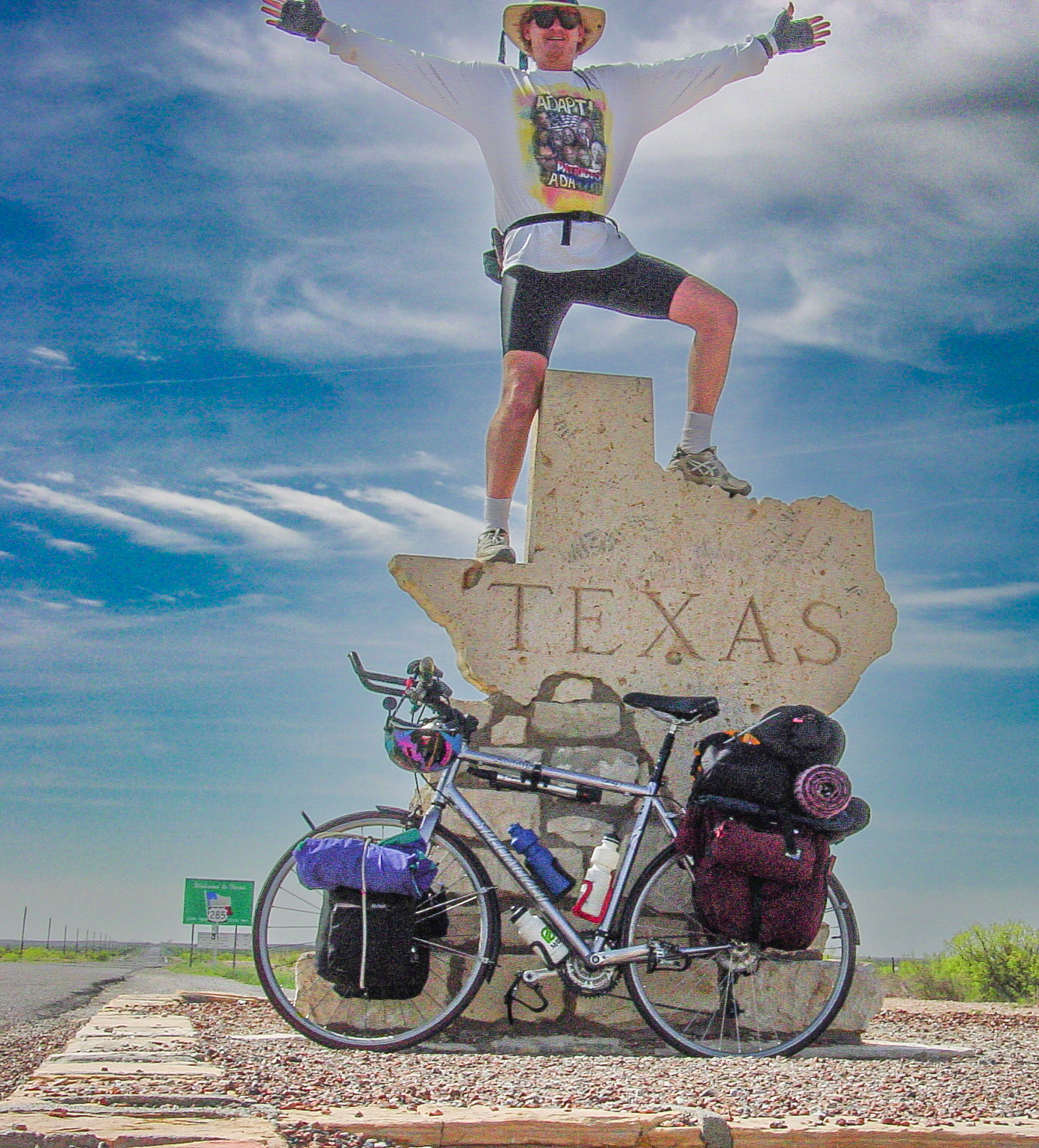 Man stands atop the Texas state line marker in the shape of the state, bicycle leans on the sign below. Blue sky with clouds in open country. The top of the man's hat is not visible.
