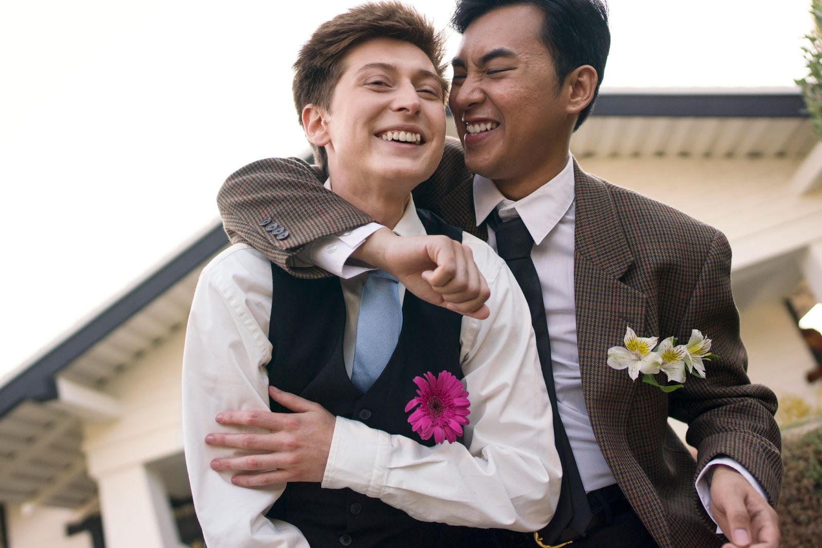Imagem de dois homens sorrindo em trajes formais. Um homem com traços asiáticos abraça o pescoço de um homem branco que está à sua frente.