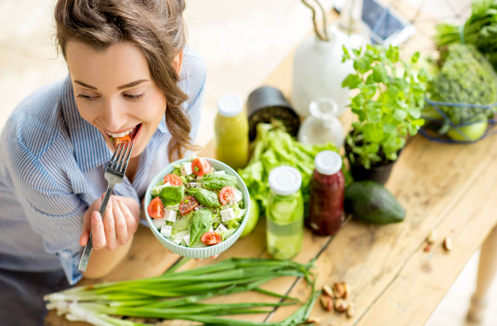A young woman, smiling and eating healthy salad sitting on the table in the kitchen.