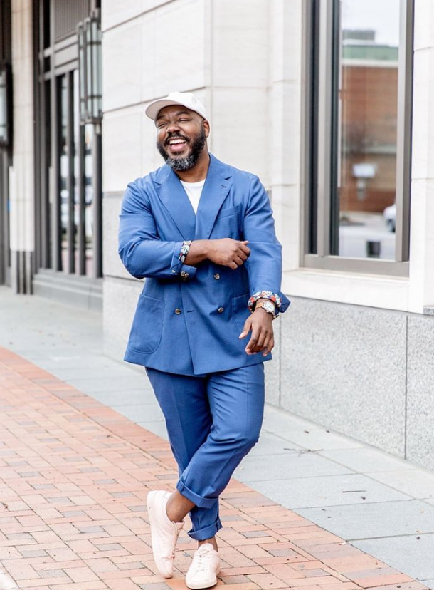 A plus size man laughs at the camera. He is wearing a blue suit and white sneakers