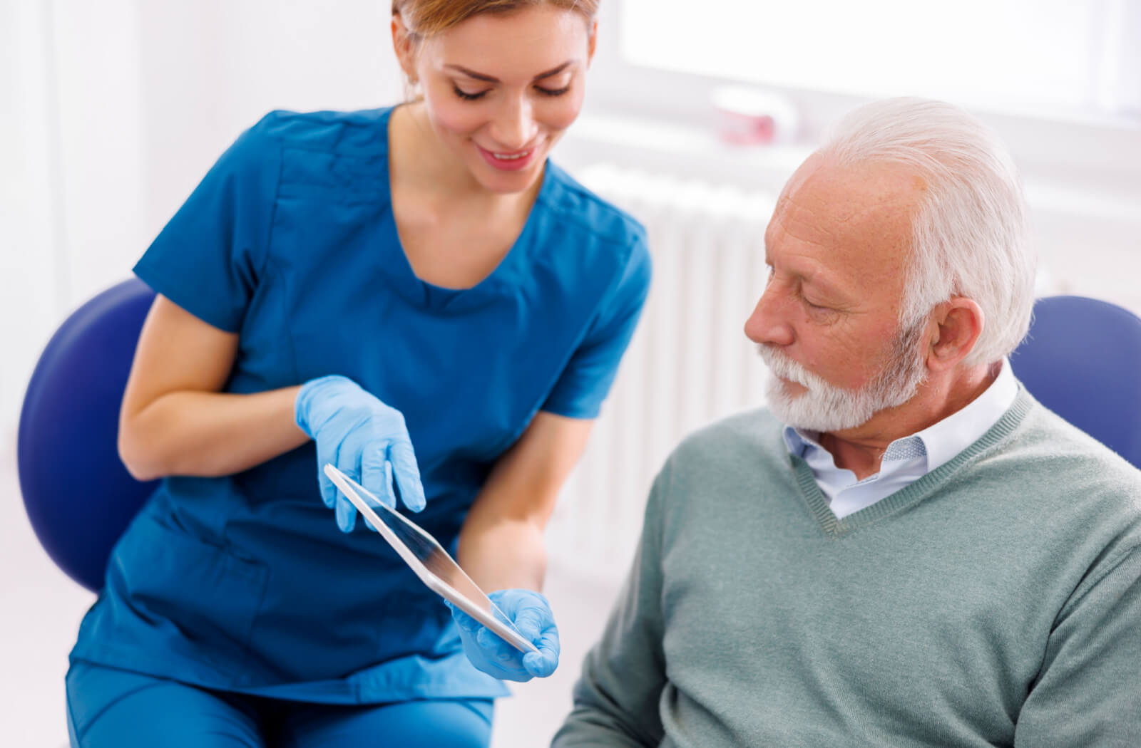 A female dentist in blue scrubs and a senior man sitting in a dentist's chair looking at a tablet.