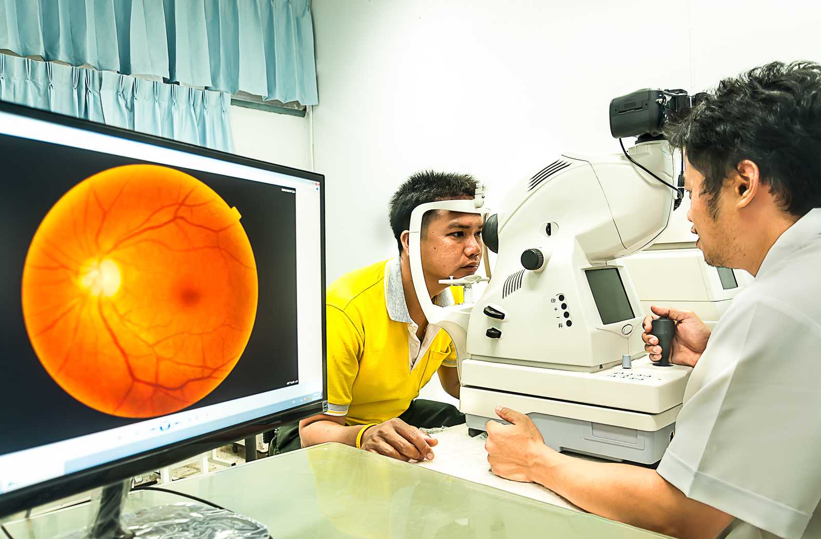 male optometry patient getting fundus photography on left eye while the retinal image renders on a nearby PC monitor