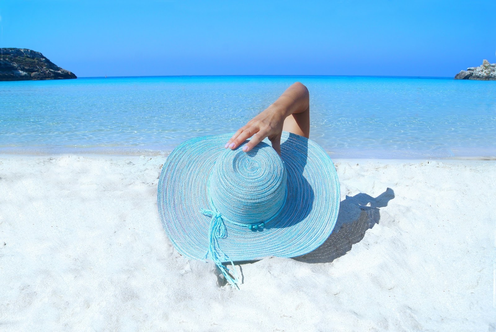 a woman lying on a white sandy beach facing clear blue water, with a sun hat getting a base tan