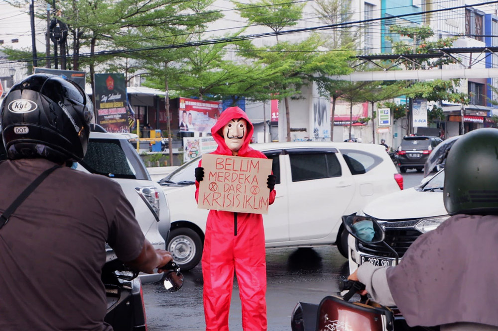 A rebel in red overalls and a Dali mask holds a sign in busy traffic.