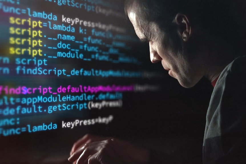 A man sits close looking at computer code on a screen, the room is dark