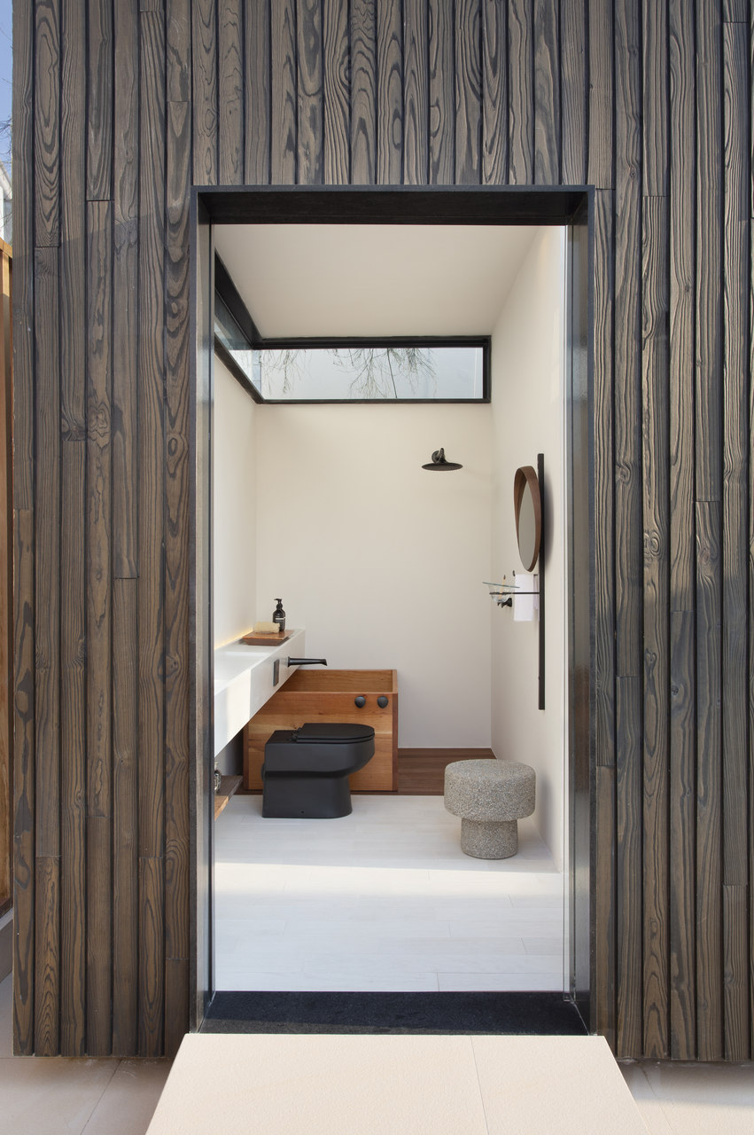 Banheiro com fachada de madeira, piso e paredes brancas, vaso sanitário e acessório pretos e banquetinha cinza 