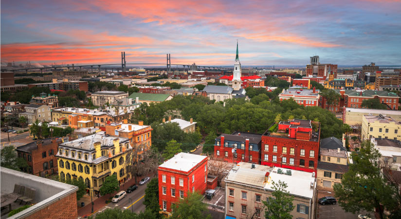 Aerial view of Downtown Savannah, Georgia, at dusk. The sky is streaked with pinks and blues and the tops of buildings poke out from above lush trees.