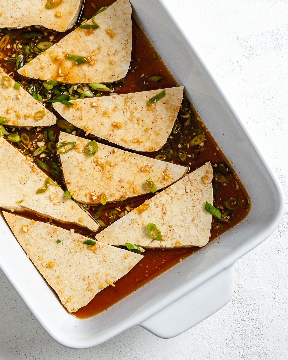 Process shot of tofu triangles in marinade in white tray.