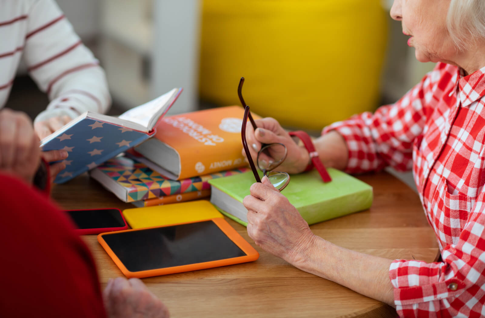 Photo of 3 seniors sitting around a table, with books and tablets in the middle. one senior is holding a pair of glasses and speaking.