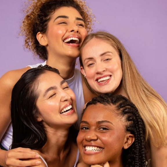 #DescriçãoDaImagem: fundo lilás com quatro mulheres diferentes entre si, sorridentes se abraçando. Foto: Divulgação Beauty UAU. 