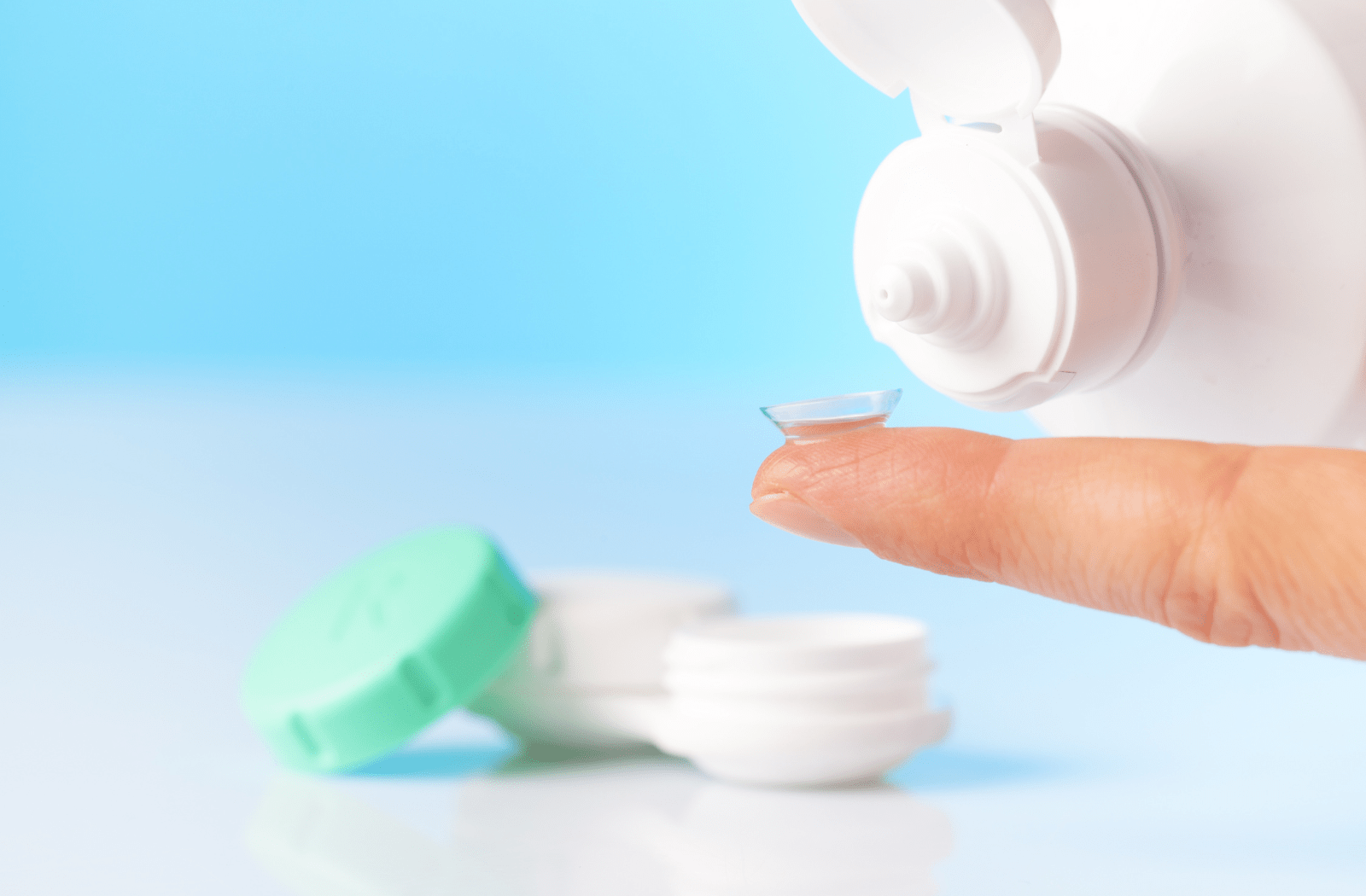 A person holding a contact lens on their index finger and they are pouring contact lens solution onto the contact with the case in the background