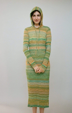Modelka ubrana w sukienkę z kapturem w cienkie rozmyte poziome paski w kolorze zielonym i pomarańczowym.