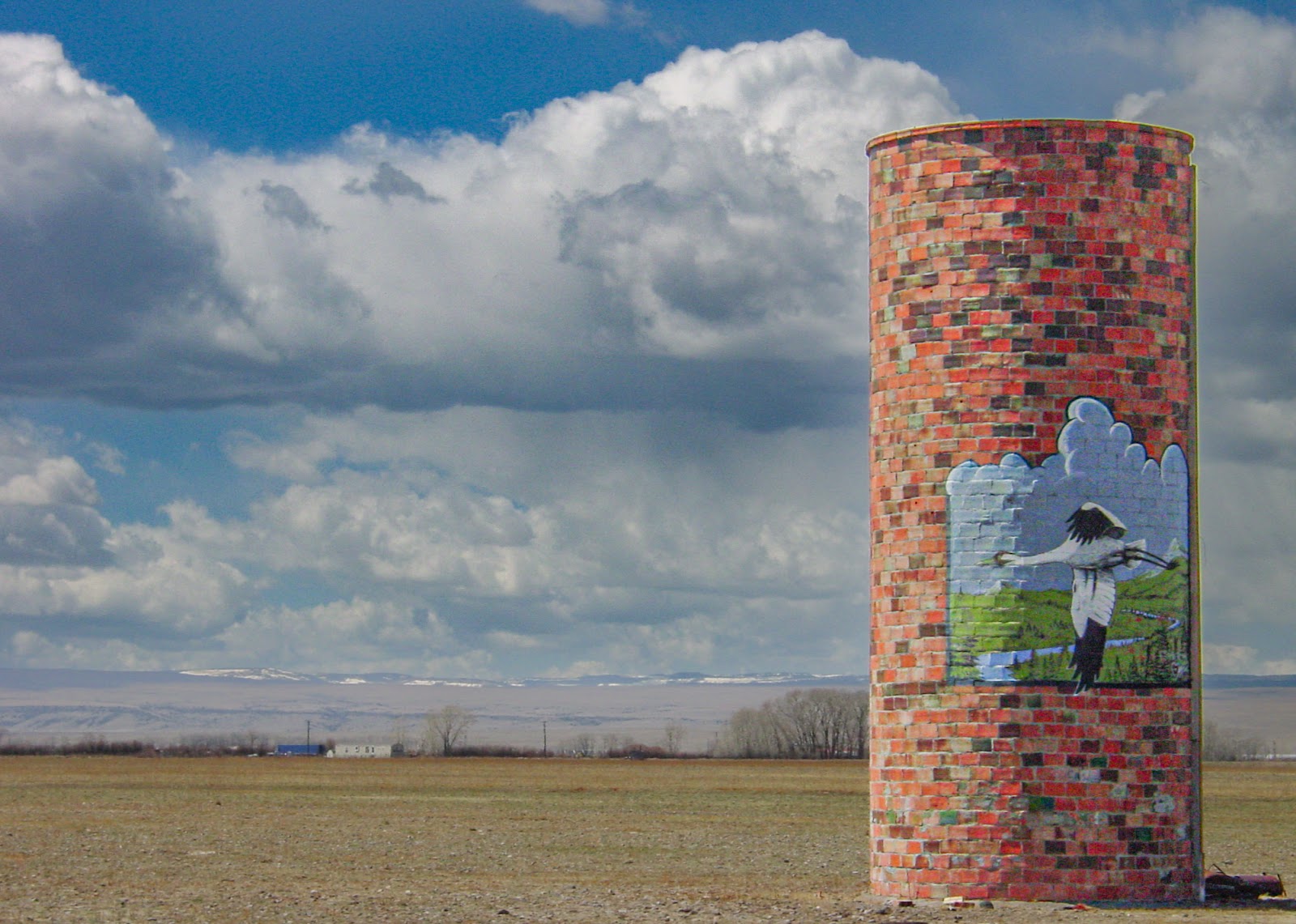 A multi-colored brik silo with mural of a woman gathering a harvest against a cloudy sky.