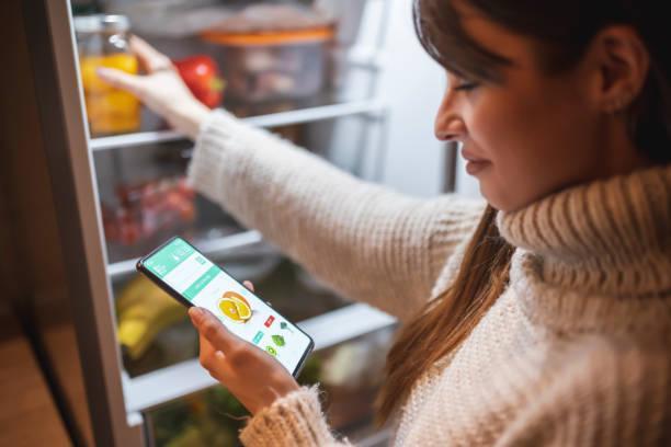 Woman ordering groceries online for home delivery Beautiful young woman standing next to an opened refrigerator door, holding a smart phone and ordering fresh fruit and vegetables online for home delivery weight loss apps stock pictures, royalty-free photos & images