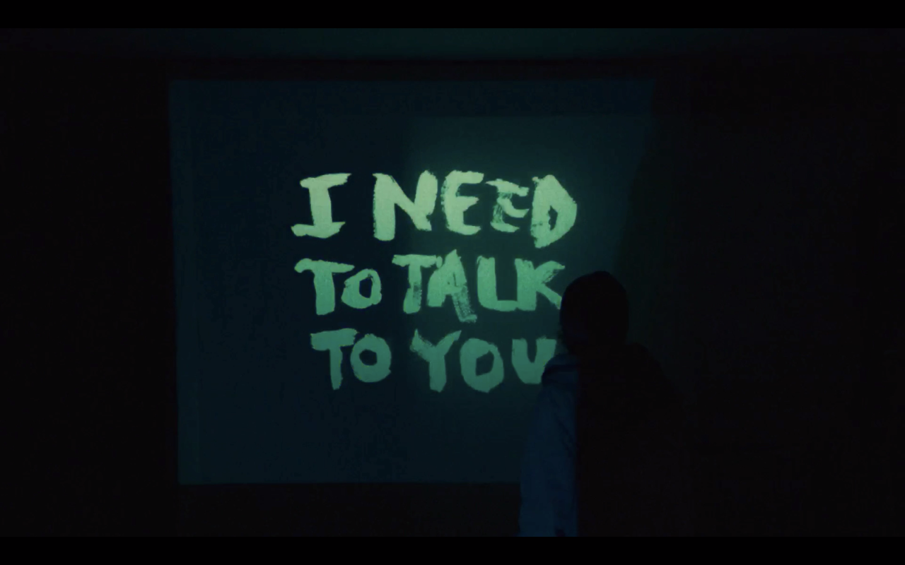 A still from We're All Going to the World's Fair. Casey stands in a dark room as an image is projected on the wall in front of her. It is a message in neon green script, saying in all caps, "I need to talk to you." 