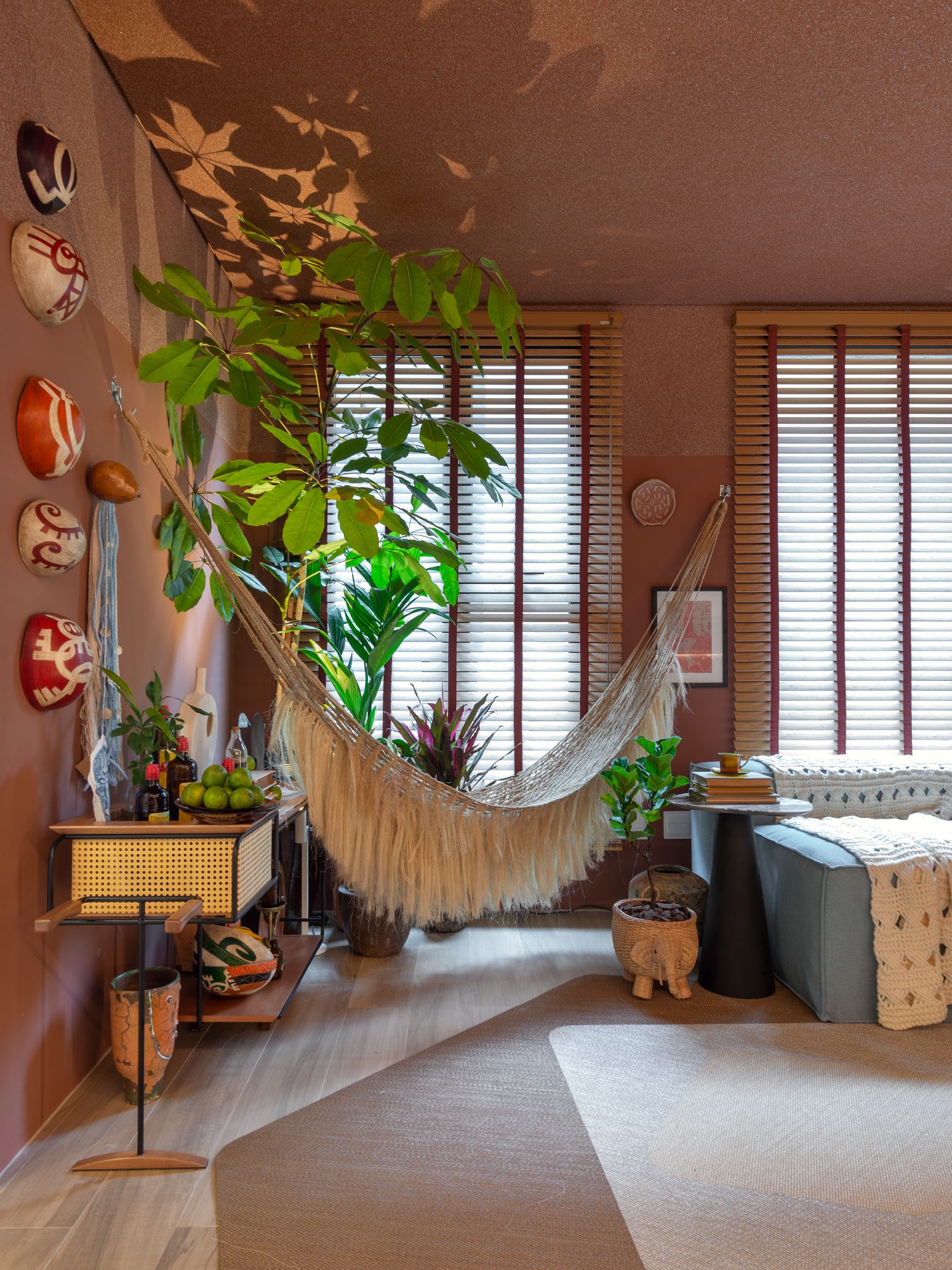 Ambiente com parede roses, piso de madeira, rede cor nude, prateleira suspensa, objetos decorativos na parede e vasos de planta.