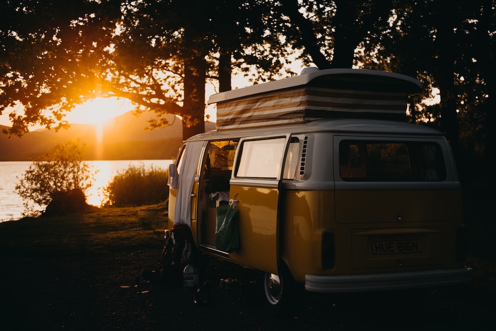 A brown van sitting under a tree, in front of a river during sunset.