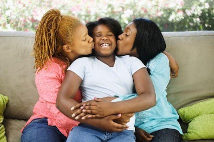 [Image is a photo of three people sitting on a couch, embracing. The person in the middle is laughing while being kissed on the cheek by both people on either side of them. The person on the left has orange locs, medium skin tone, and a pink shirt. The person in the middle has black hair, dark skin and a plain white T-shirt. The person on the right has straight black hair, dark skin, and a blue shirt.]