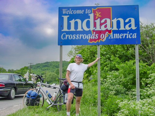 A cyclist stands by the Indian state line sign that says: Welcome to Indiana, crossroads of America.