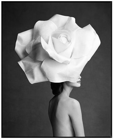 Fotografia em preto e branco, modelo está de perfil, cabelos presos e pretos, aspecto sedoso. Usa um chapéu que cobre parte do seu rosto. Deixando visível apenas a boca. O chapéu que usa parece um grande e belo botão de rosas brancas.