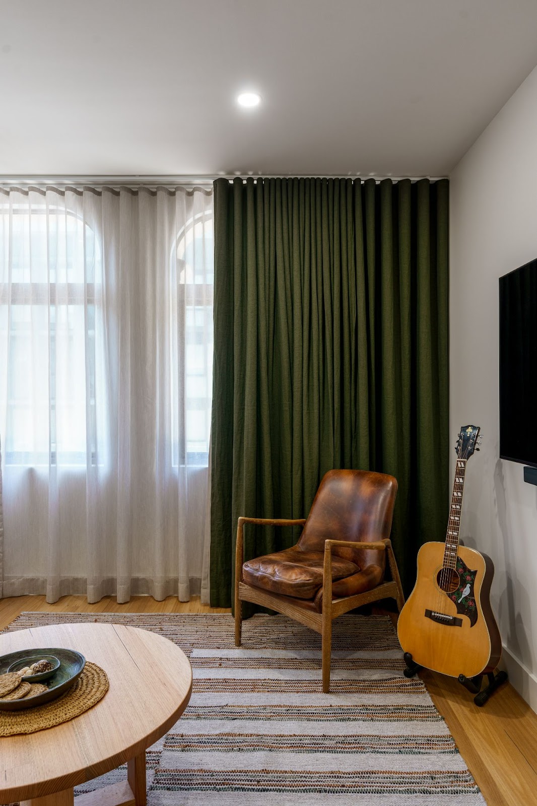 A cozy corner in a living room featuring a leather armchair beside a classic guitar resting against the wall. The space is softened by rich green curtains, sheer drapes, and a round wooden coffee table with rustic decor.