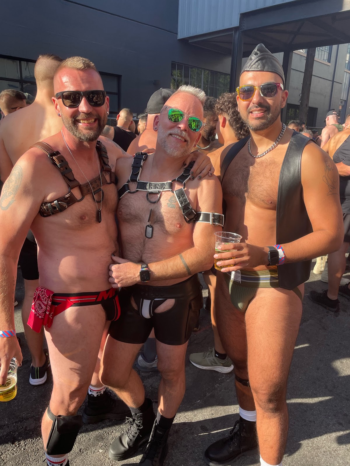 Three gay muscle daddies wearing jockstraps, leather harnesses and leather vests outside and drinking during Folsom street fair 2023 in San Francisco