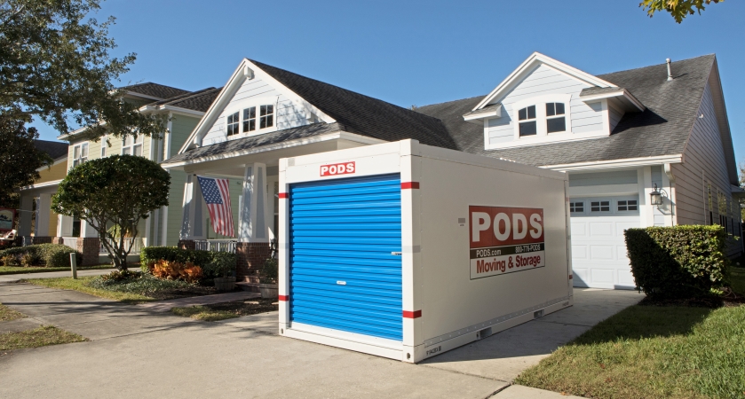 A PODS moving and storage container sits in the driveway of a white house with an American flag. The signature blue door is facing the street side.