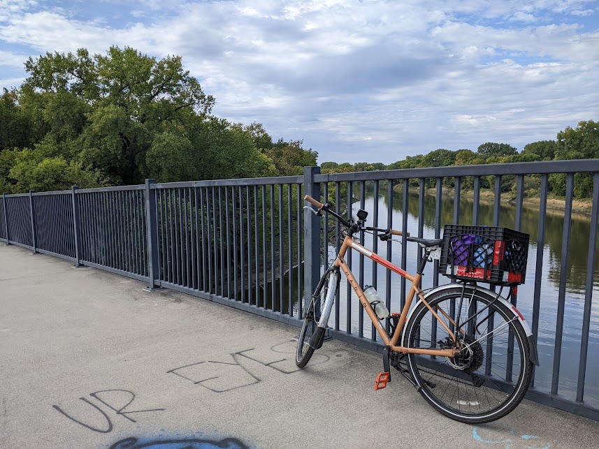 Tan bicycle with rack, fenders, and milk crate leaning against a black painted metal railing on a slightly graffiti covered concrete bridge with a tree lined river in the background. 